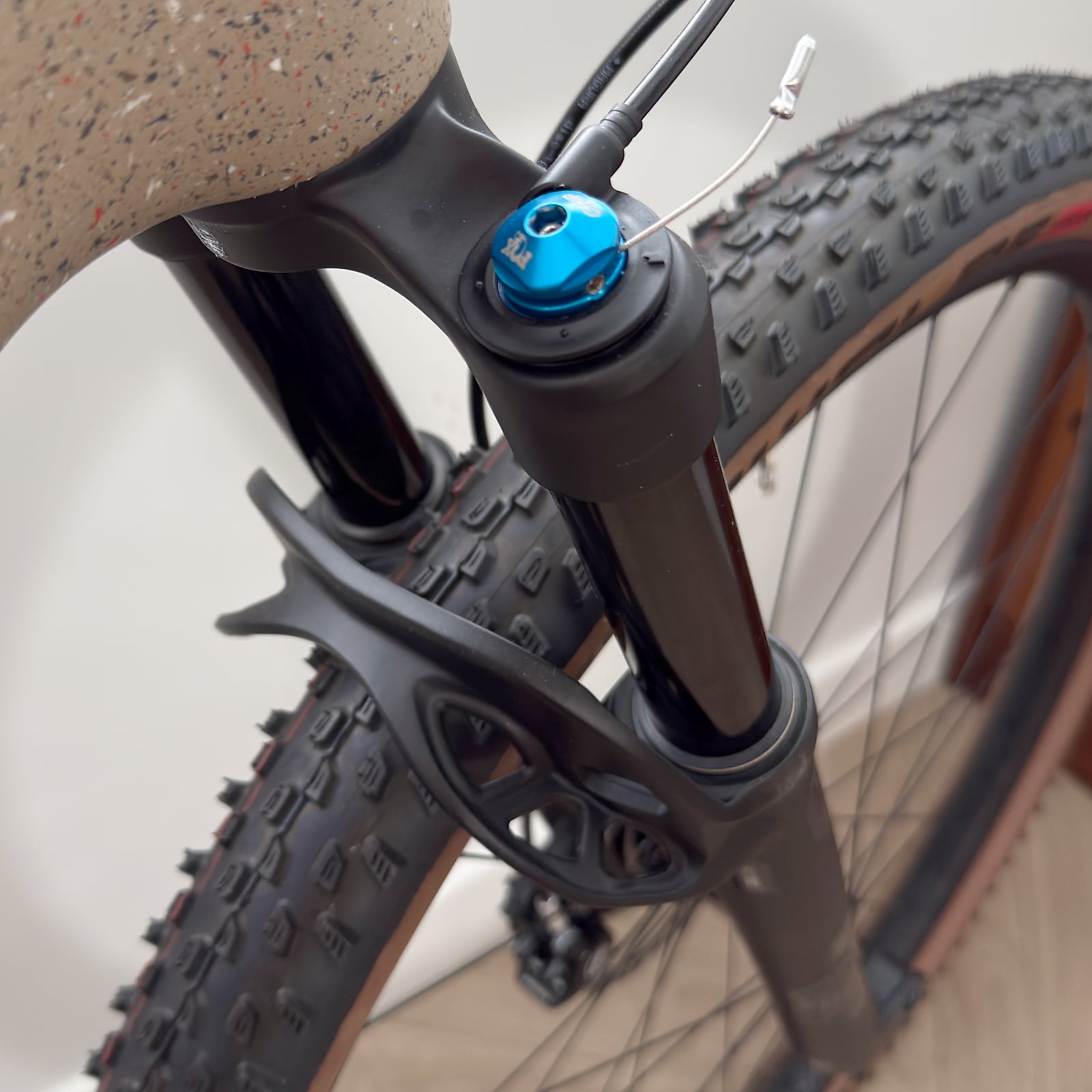 Close-up of blue trigger shifter on black mountain bike handlebar, with knobby tyres and frame visible, indoor setting.