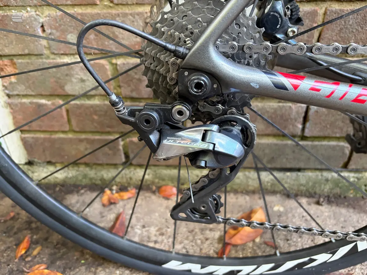 Shimano rear derailleur and cassette on a red and grey mountain bike, drive side close-up against a brick wall.