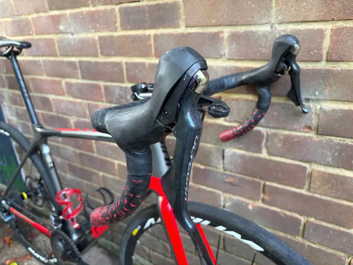 Close-up of Shimano drop bar shifters and brake levers on a black and red road bike, non-drive side, against a brick wall.