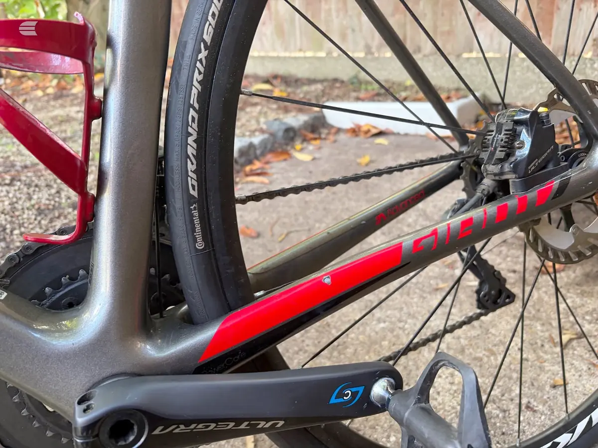 Red and grey road bike, drive side close-up showing rear derailleur, chain, cassette, and seat tube branding, on pavement with autumn leaves.