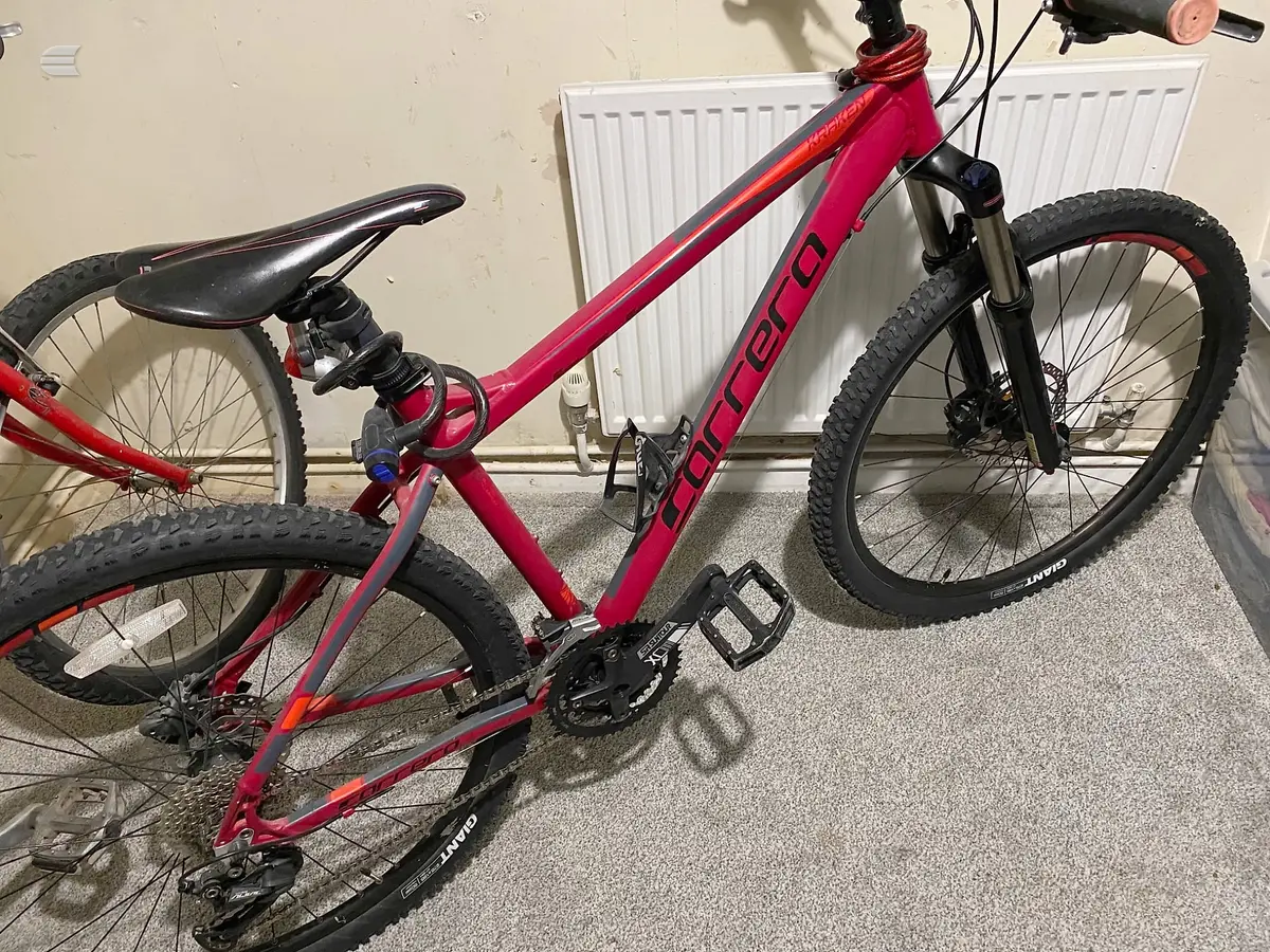 Red Carrera mountain bike, non-drive side, indoors against a white radiator and beige wall.
