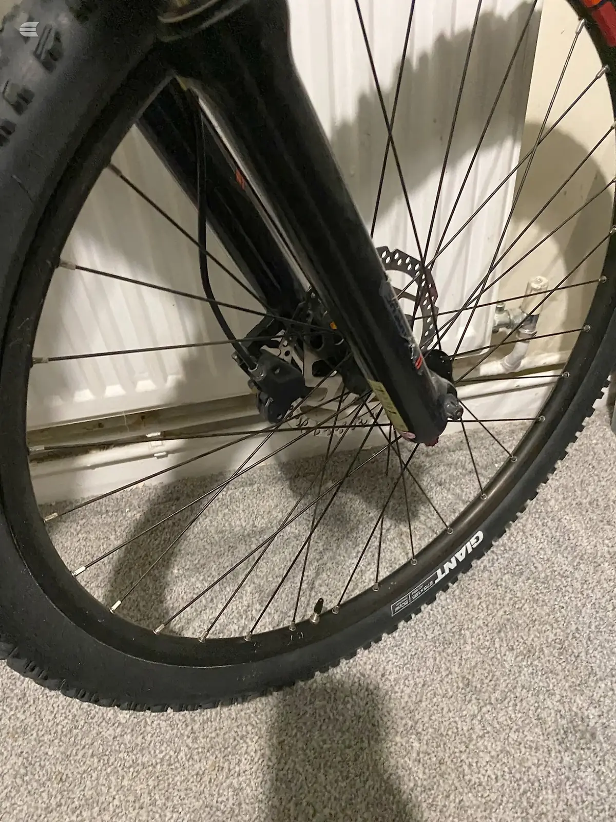Black mountain bike front wheel with disc brake, close-up on drive side, indoors on grey carpet with white blinds in background.