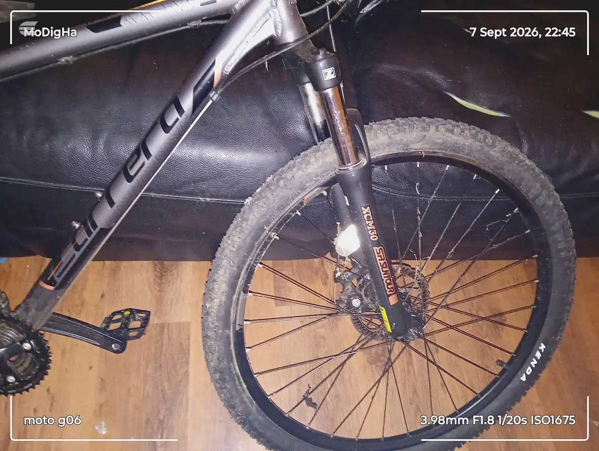 Front wheel of black hardtail mountain bike on wooden floor, drive side, close-up showing tread and disc brake.