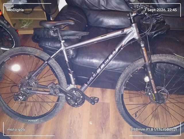 Silver hardtail mountain bike with disc brakes, dark saddle and grips, on wooden floor indoors, drive side view, partially obscured by person's legs wearing