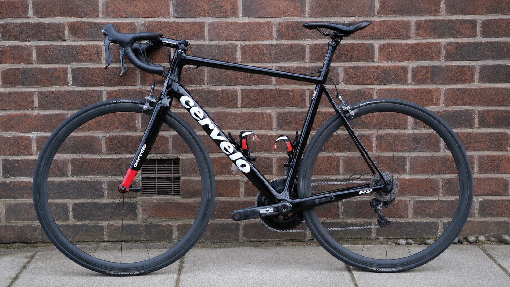 Black Cervélo R2 road bike with red accents, drive side, leaning against a red brick wall on pavement.