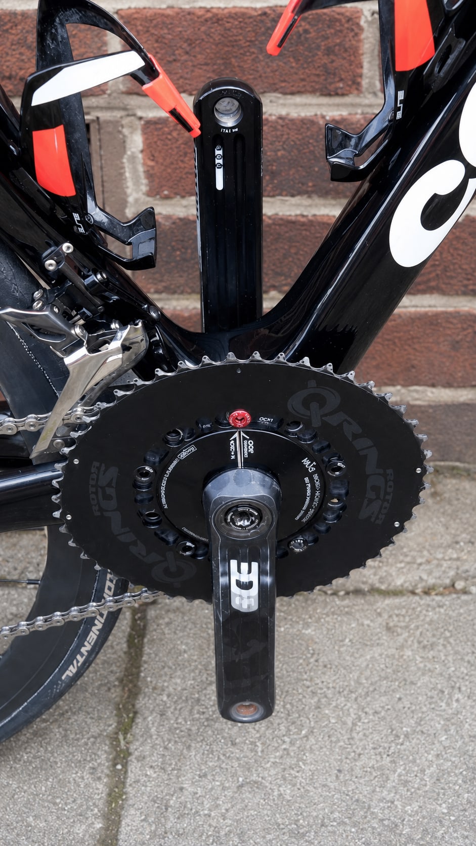 Close-up of black crankset and chainring on a black and red bike, drive side, against a brick wall.