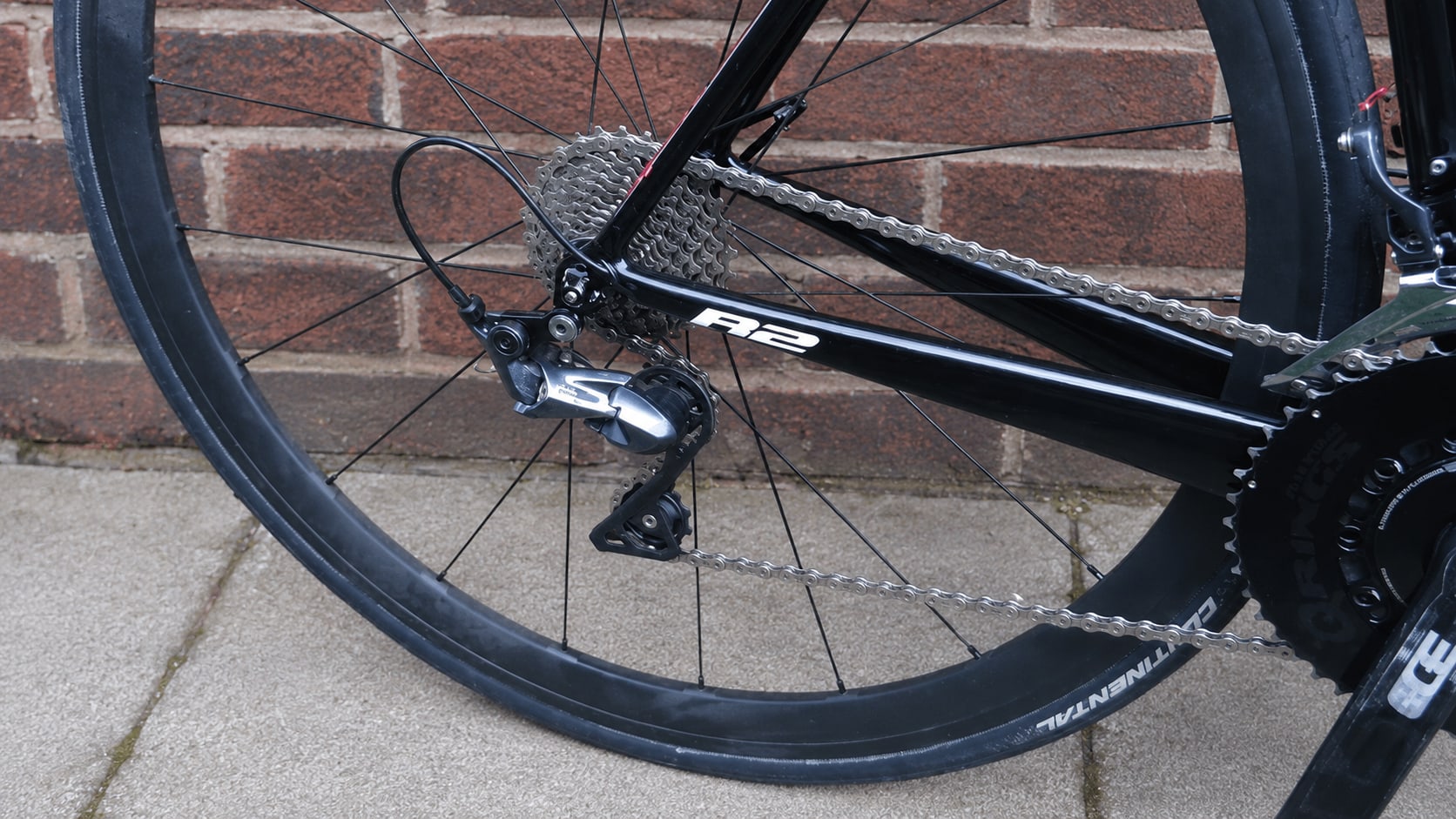 Close-up of bicycle drivetrain components against brick wall: rear derailleur, cassette, and chain on black road bike, drive side.