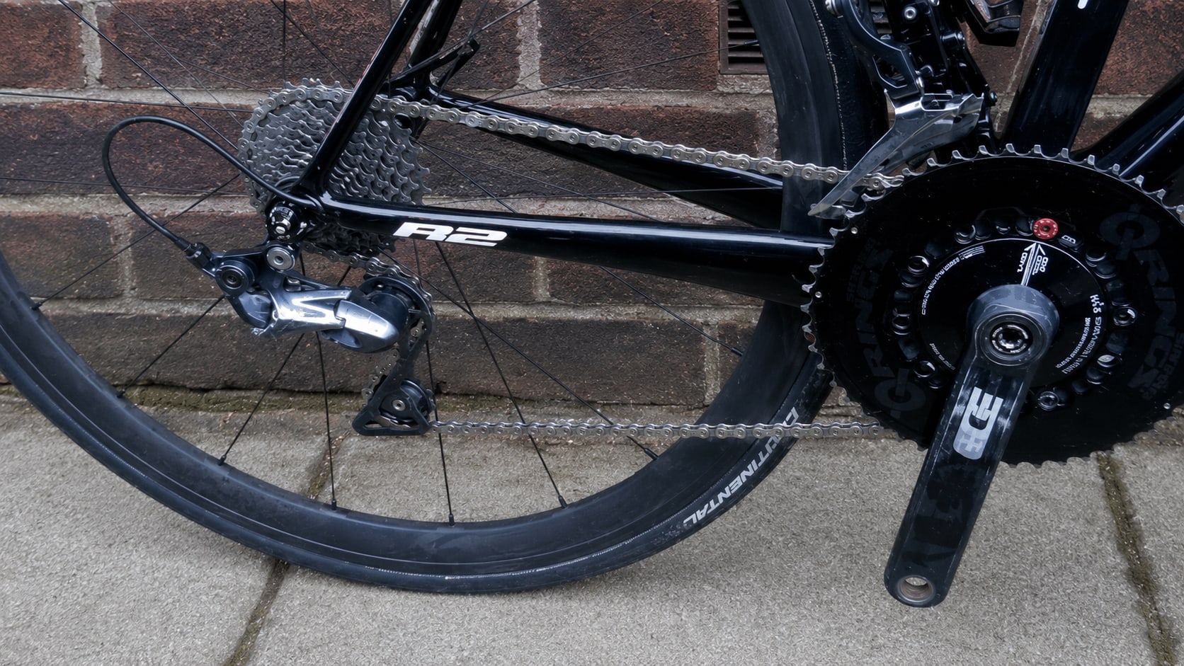 Black road bike drivetrain detail, drive side, showing rear derailleur, cassette, chain, and crank against brick wall background.
