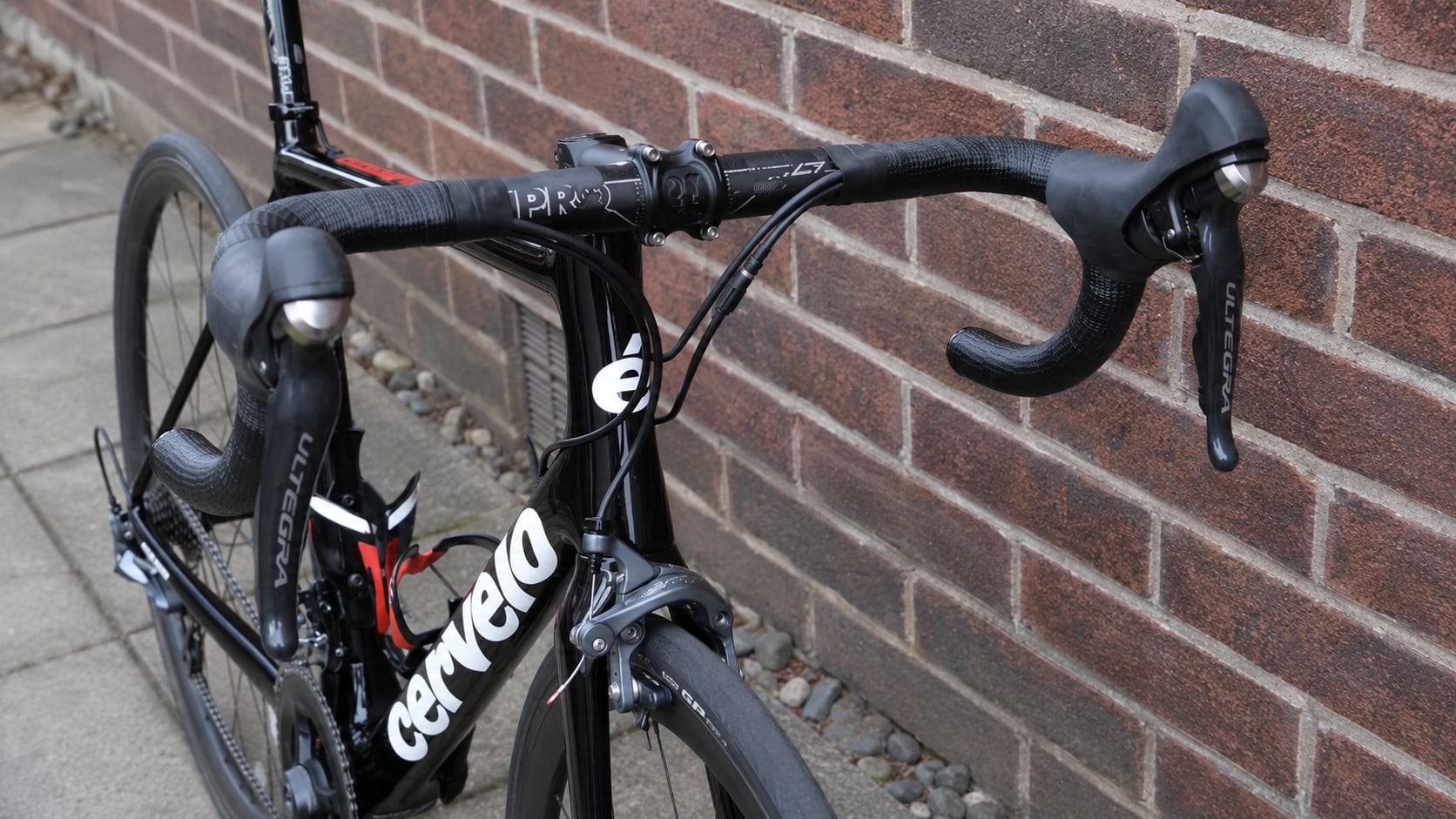 Black Cervélo road bike with drop bars and wrapped handlebar tape, non-drive side against a red brick wall, front wheel visible on left.