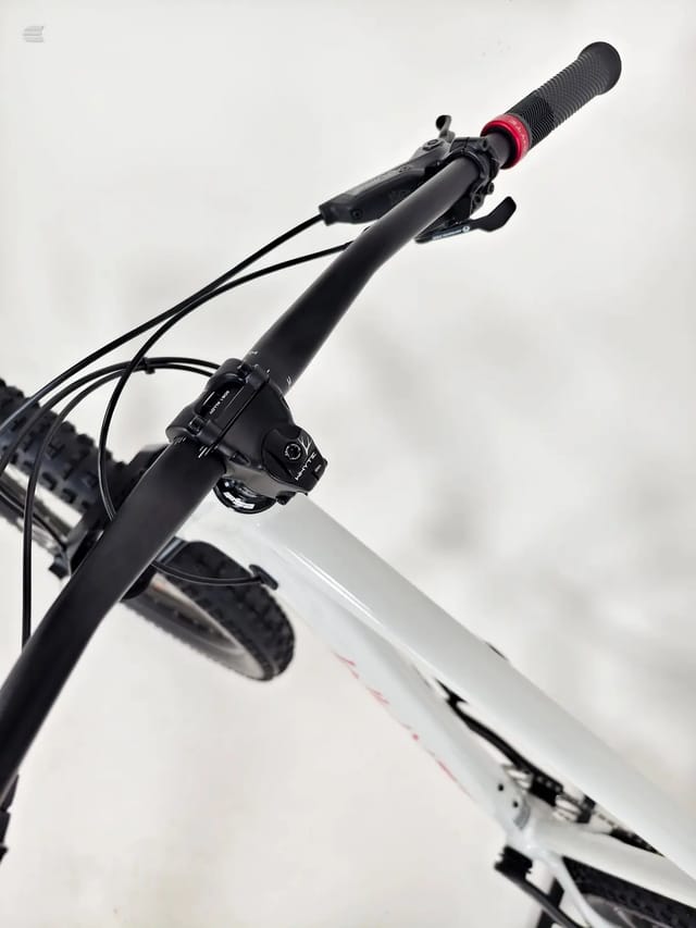 Close-up of black mountain bike handlebars and stem with red grip, angled upward against white sky, showing brake levers and cables.