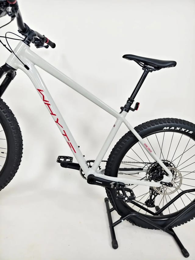 White Maxxis hardtail mountain bike with red accents, drive side, on a black stand against a white background.