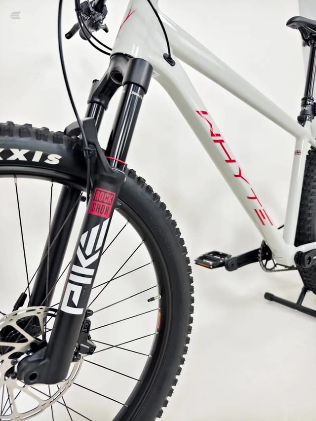 White and black hardtail mountain bike on a stand, drive side, against a plain white background. RockShox fork and Axis tyres visible.