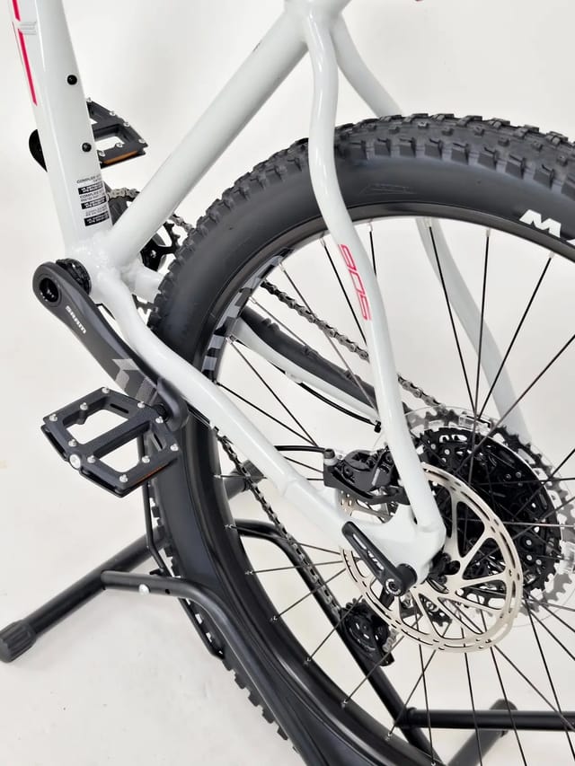 White hardtail mountain bike on a black stand, drive side, against a white background. Close-up angled view showing front fork, both wheels, and lower frame.
