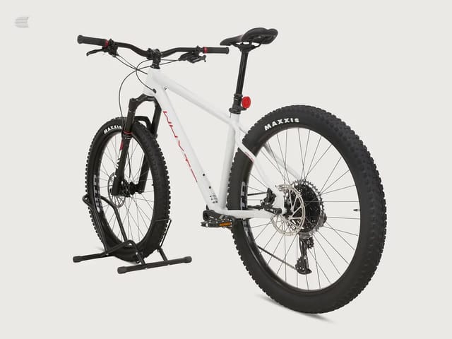 White hardtail mountain bike with black Maxxis tyres, displayed on a stand against a neutral background, three-quarter view showing drive side.