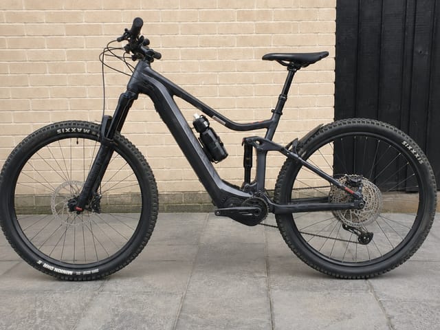 Black full-suspension electric mountain bike on paved ground, drive side, against a brick wall and dark wooden door.