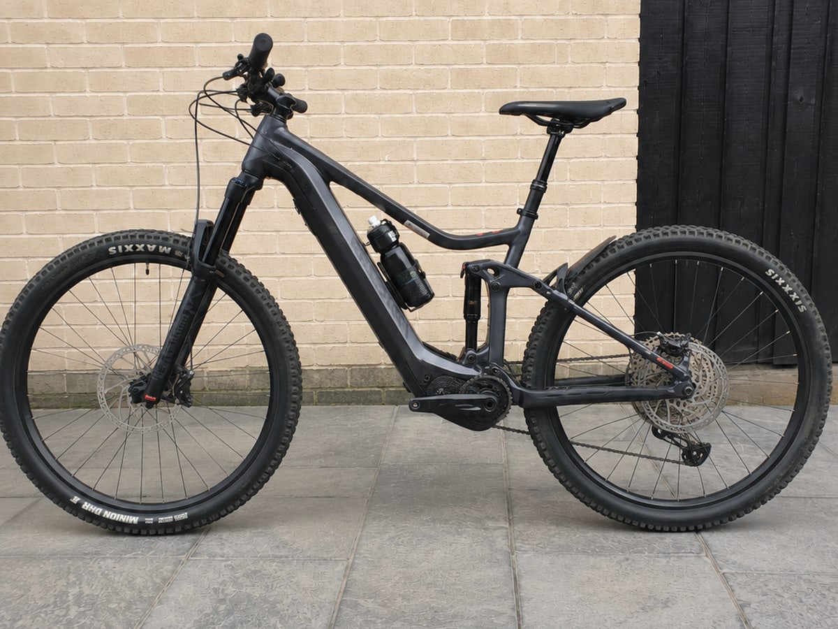 Black full-suspension electric mountain bike on paved ground, drive side, against a brick wall and dark wooden door.