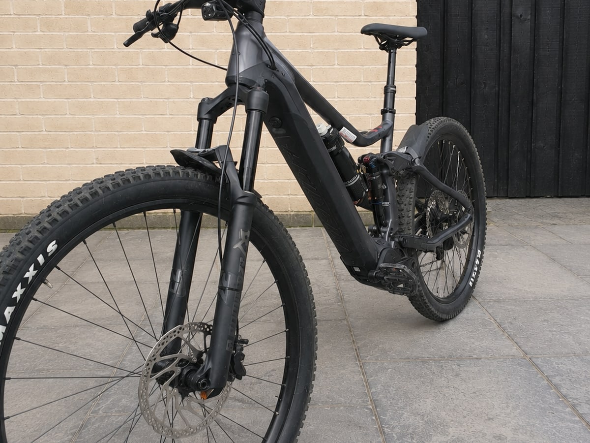 Black full-suspension electric mountain bike on paved ground, three-quarter drive-side view, against brick wall and dark shutter.