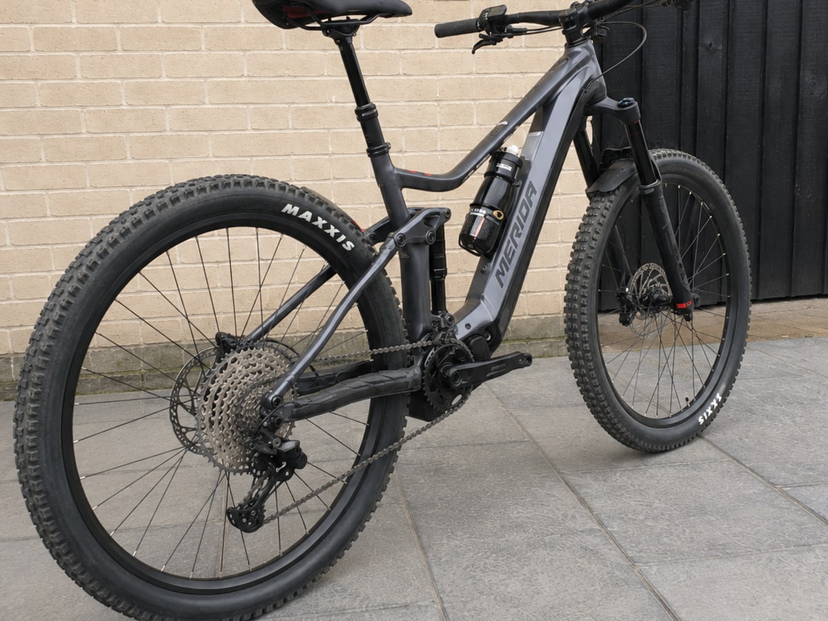 Grey full-suspension mountain bike on paved ground, drive side, against a brick wall and black door.