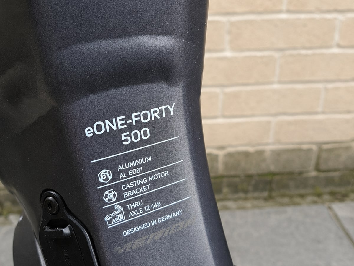Close-up of black aluminium motor bracket on eONE-FORTY 500 e-bike, showing casting motor bracket and axle specifications, against brick wall background.