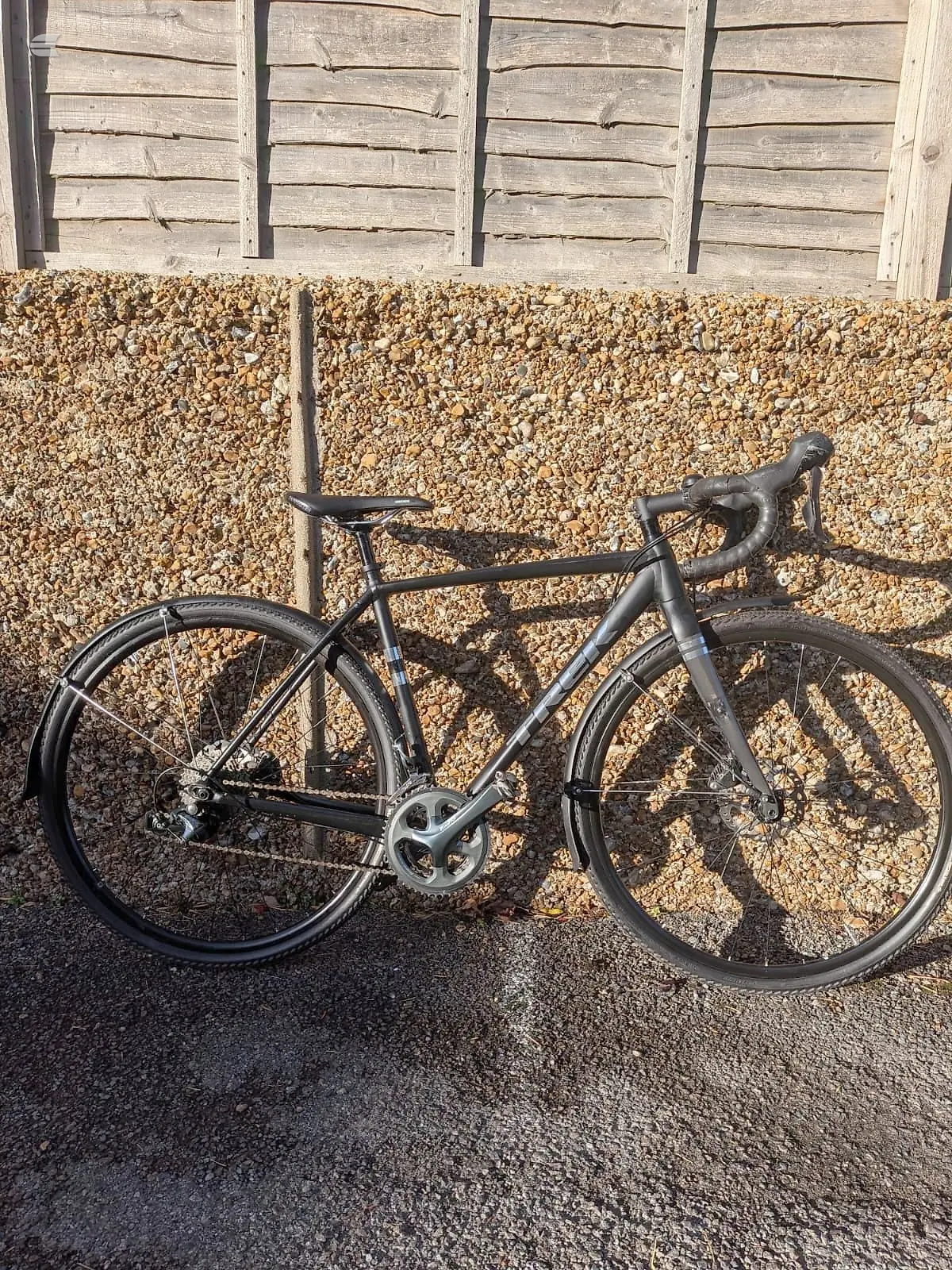 Black road bike with drop bars on gravel surface against wooden fence, drive side view.