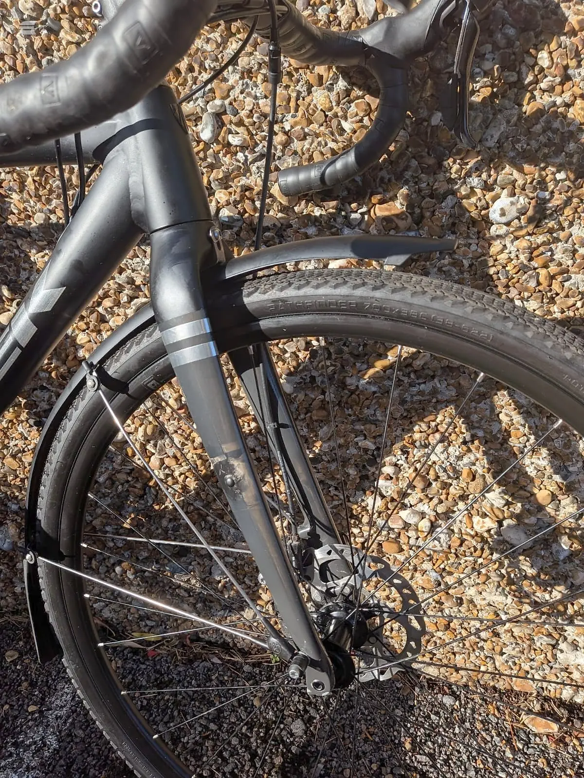 Black gravel bike on gravel ground, three-quarter view from drive side, showing frame, handlebars, front wheel and partial rear wheel.