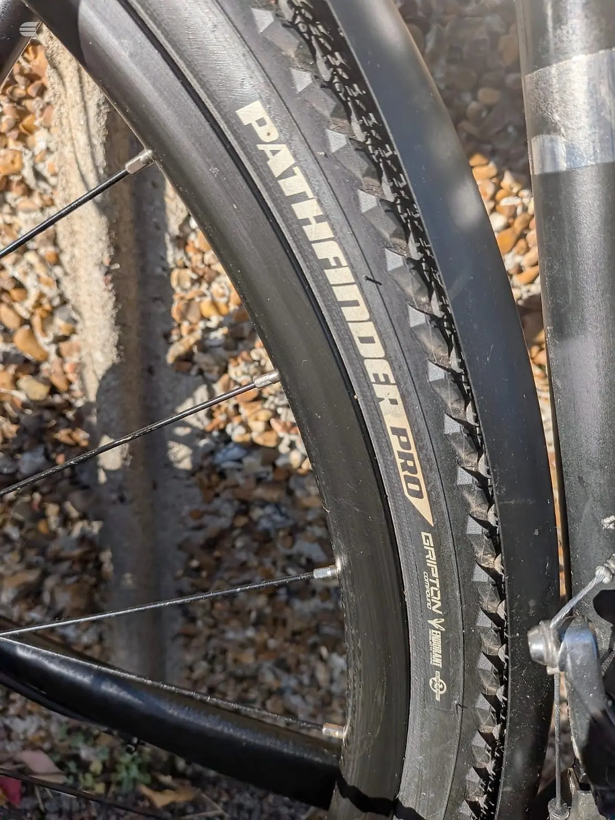 Close-up of black Schwalbe Panaracer tyre on bicycle wheel, drive side, against gravel ground with metal frame visible.