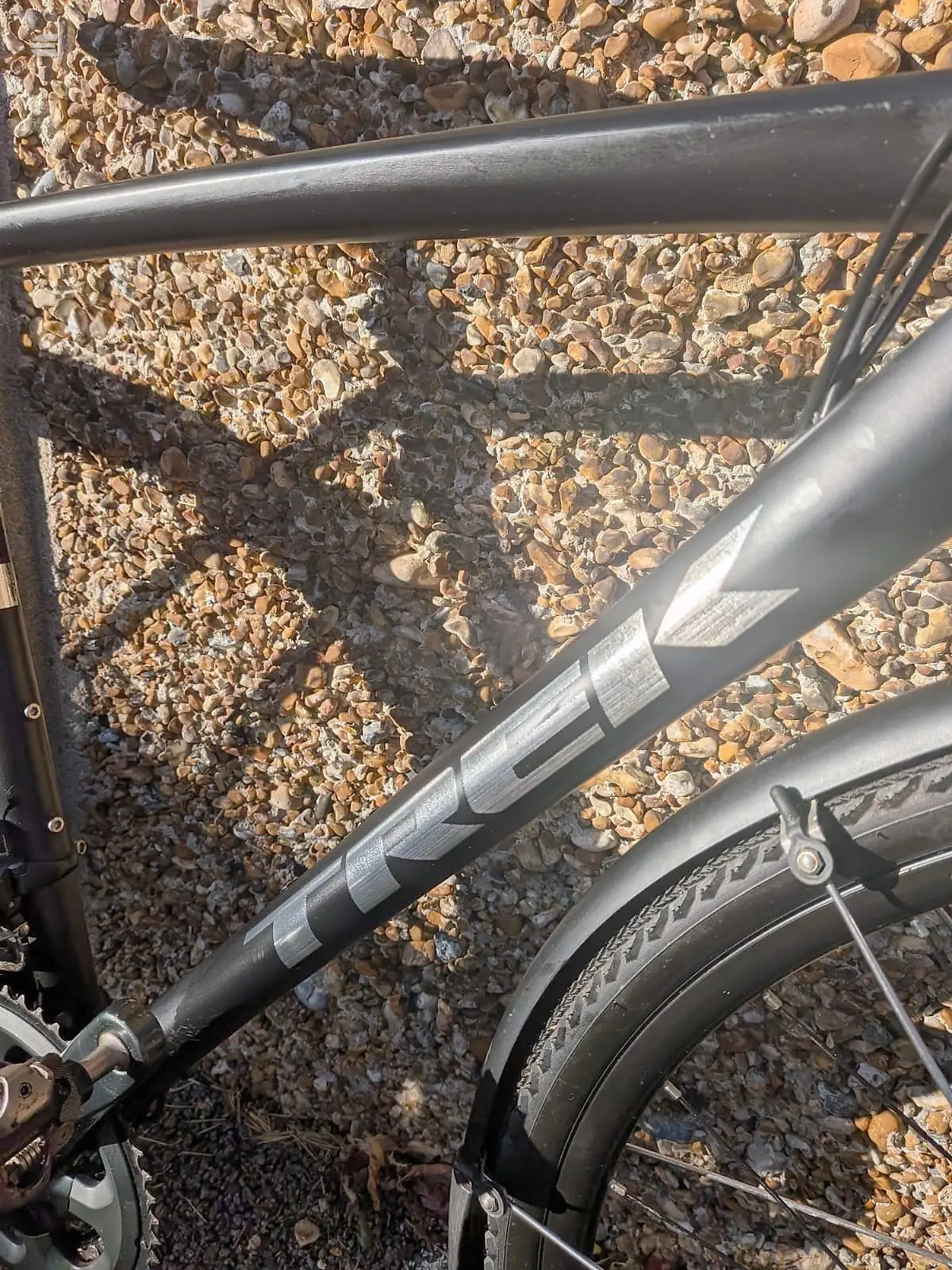 Black steel bicycle frame on gravel ground, non-drive side angle, showing seat tube and chain stays with partial wheel visible.
