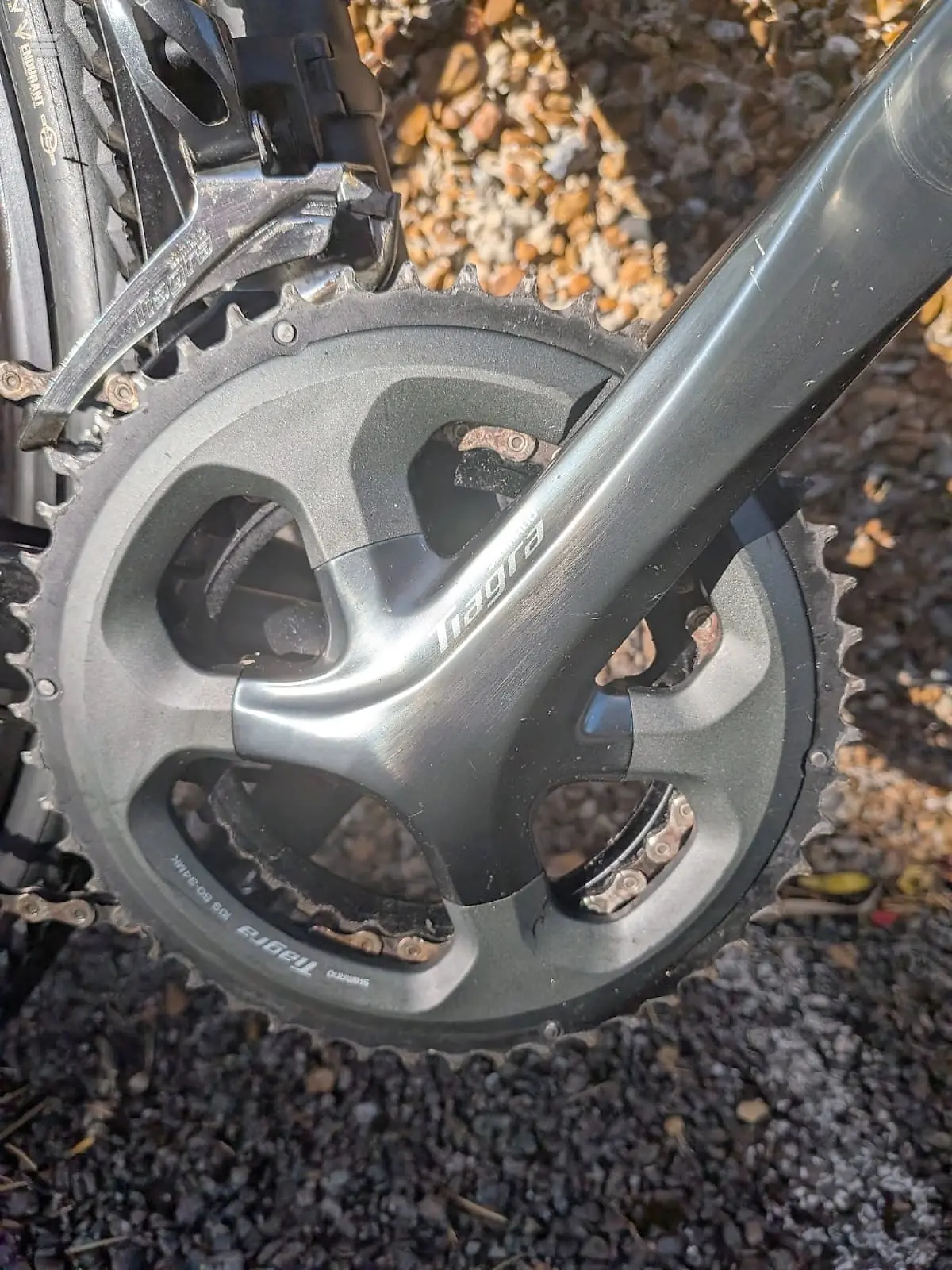 Close-up of bicycle crankset and chainrings, drive side, showing triple chainring setup with dirt and gravel background.