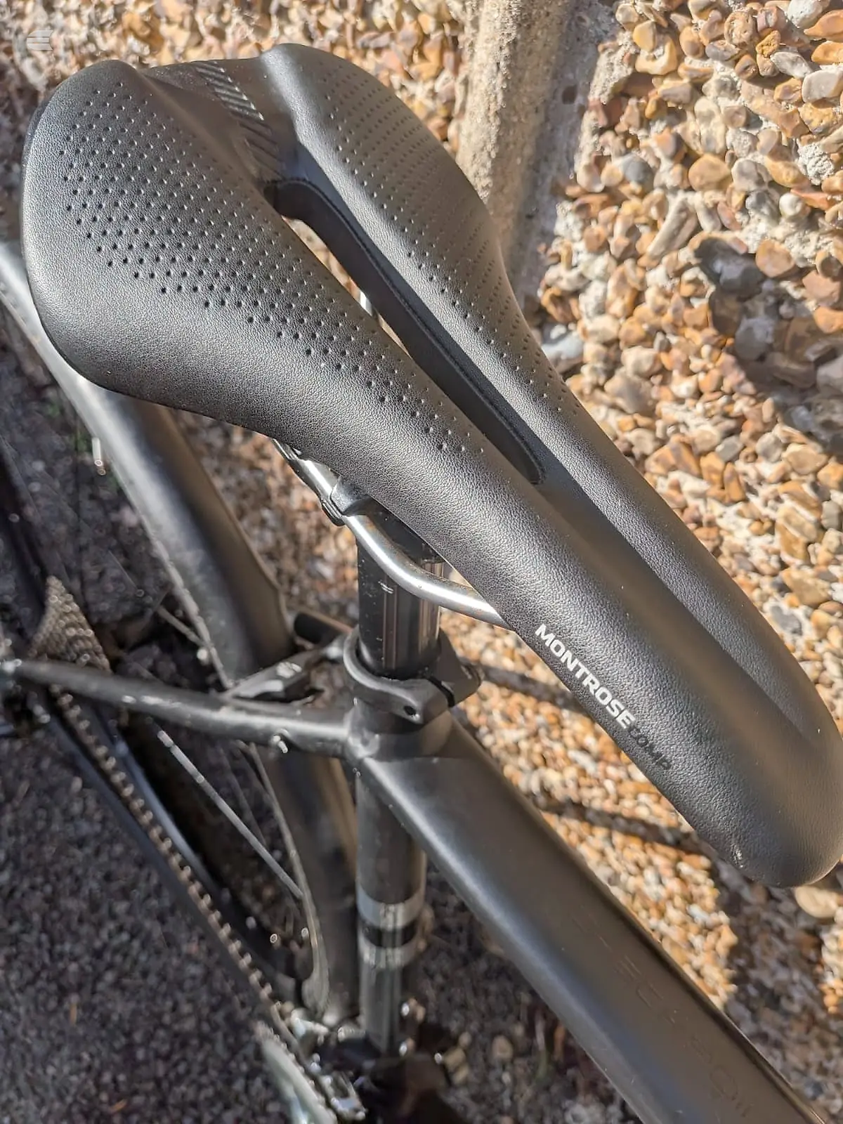 Black Montrose saddle with perforated microfiber cover on bicycle frame, mounted on gravel surface with scattered stones.