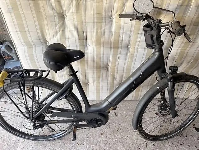 Black step-through e-bike with mudguards and rear rack, parked indoors against striped curtain, drive side view.