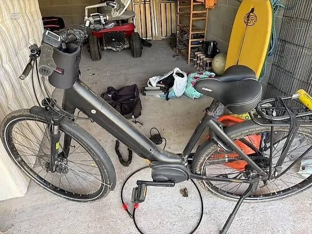 Silver step-through hybrid bike with front basket, parked indoors in a garage alongside an orange mountain bike, non-drive side view.