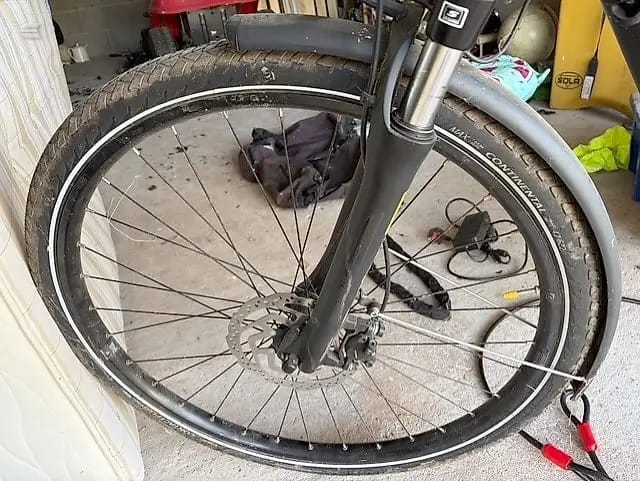 Front wheel of a grey hybrid or commuter bike on concrete garage floor, non-drive side, with frame and fork visible behind. Tyre shows wear.