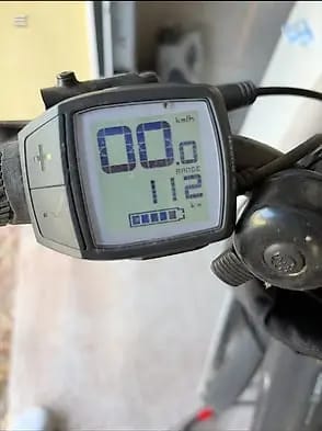 Close-up of a bicycle computer display mounted on handlebars, showing speed and distance readings, with blurred bike frame and wheel in background.