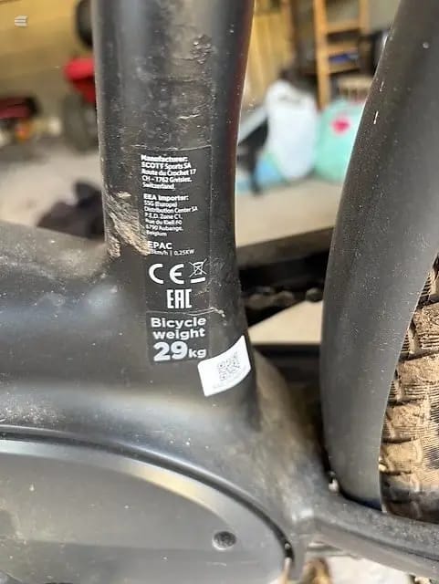 Close-up of black bicycle seat tube and frame detail showing weight specification label, CE marking, and regulatory text on dark background in workshop setting.