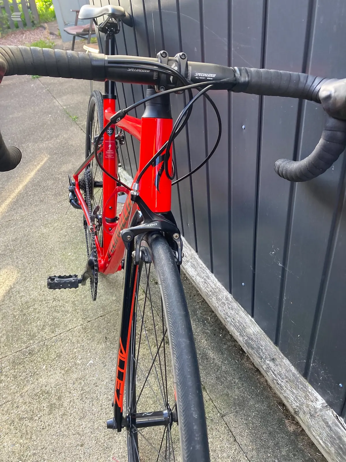 Red Specialized road bike, drive side, leaning against a dark corrugated metal shed with wooden fence rail in foreground.