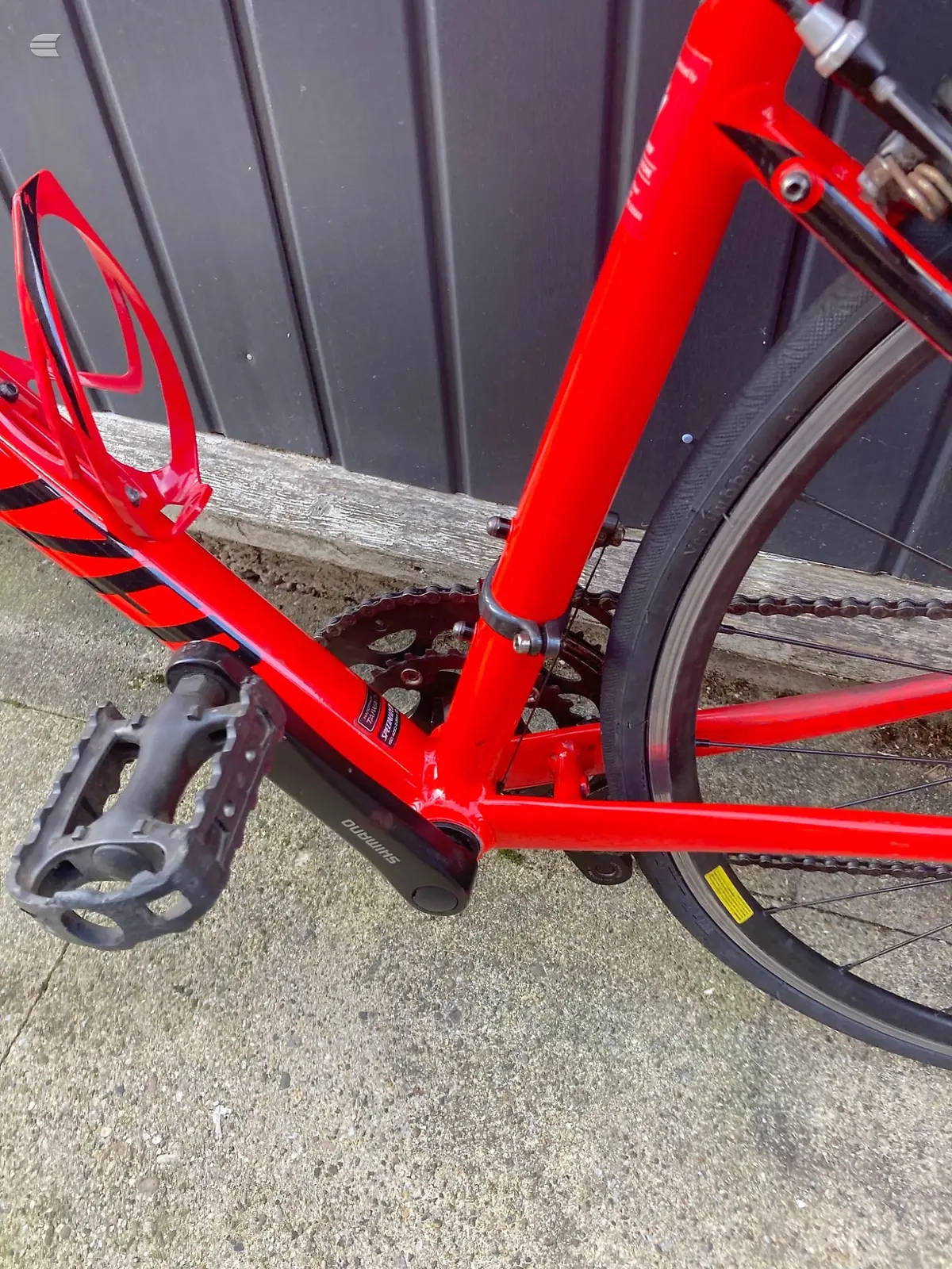 Red hybrid or commuter bike, drive-side close-up of crankset, pedals, and chain against grey corrugated metal backdrop.