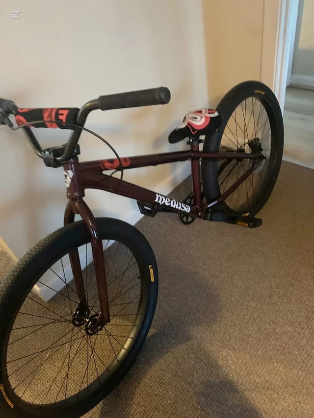 Maroon fixed-gear bike with red and black handlebar tape, viewed from the drive side at a slight angle indoors on carpet. Both wheels visible, front wheel in