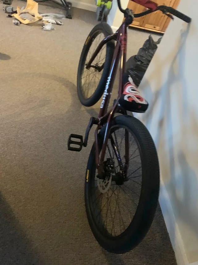 Purple and blue fixed-gear road bike indoors on carpet, non-drive side, leaning against white wall.