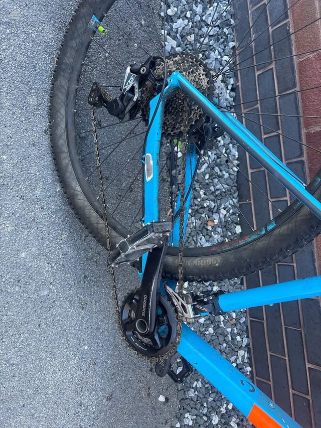 Blue road bike with orange accents, overhead view on gravel and brick paving, drive side visible with Shimano rear derailleur and cassette.