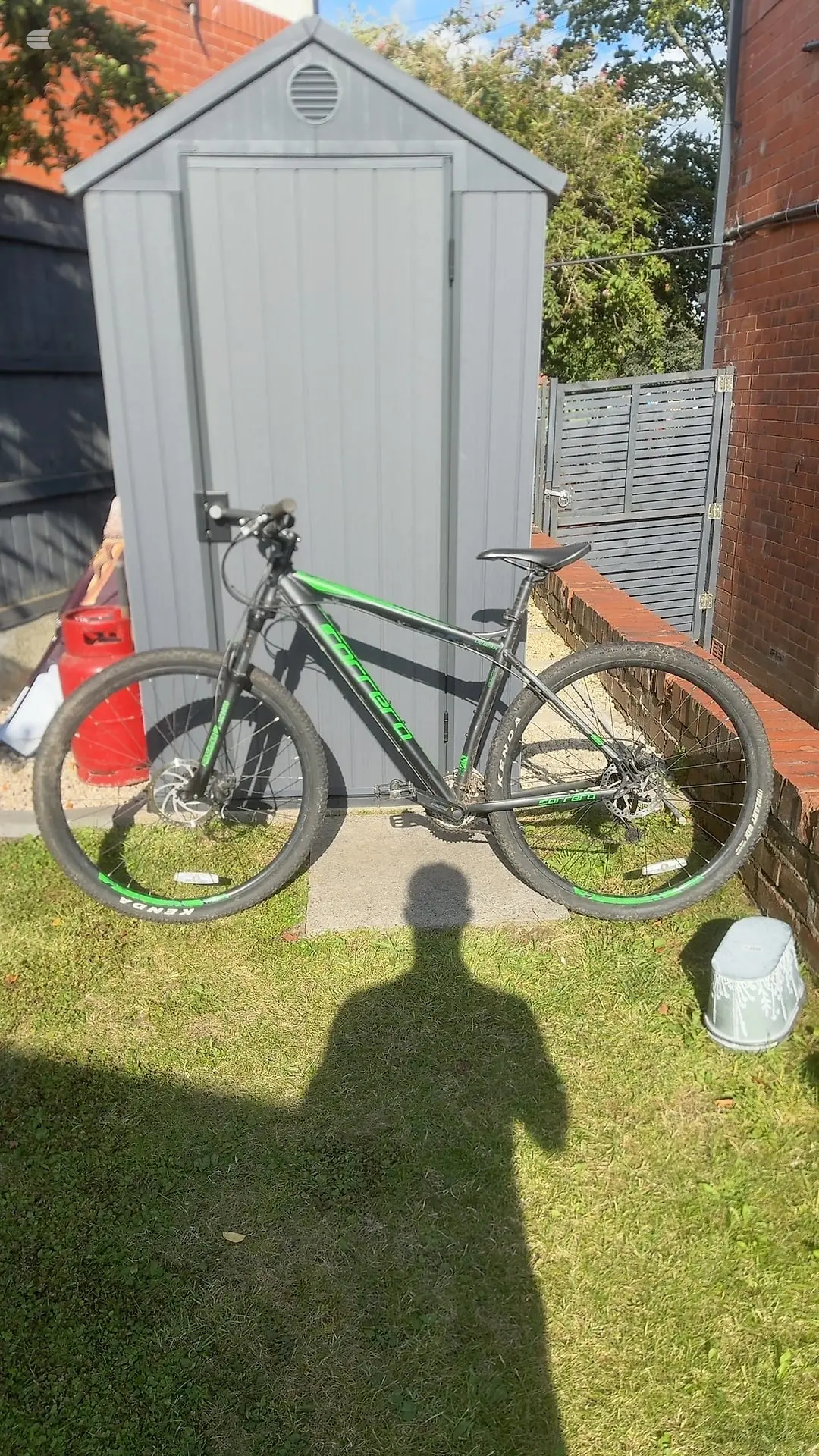 Black and green hardtail mountain bike, drive side, standing on grass in front of a grey metal storage shed in a residential garden.