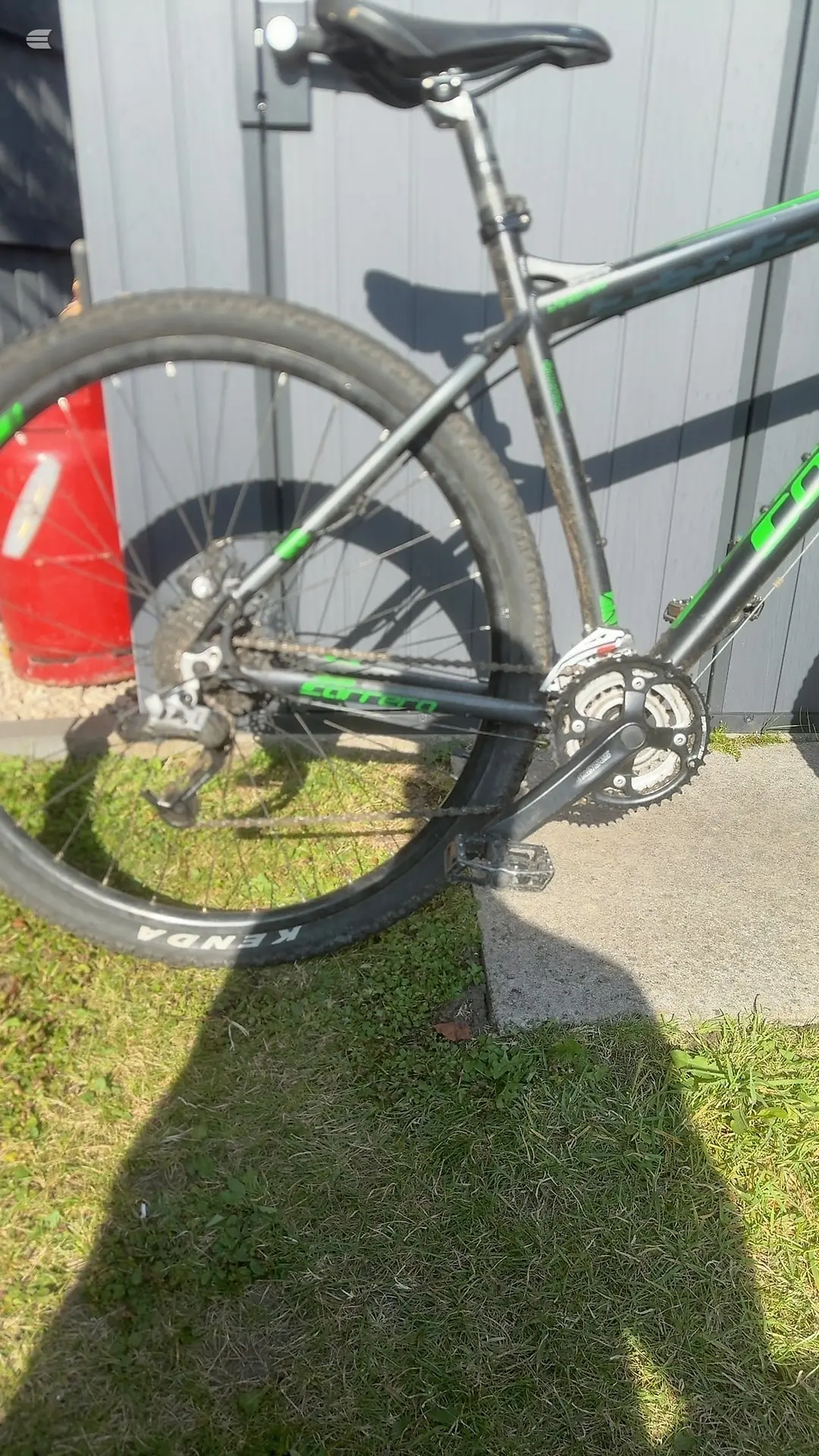 Grey and green hybrid bike with black saddle and tyres, three-quarter view from drive side, parked on grass against a metal shed.