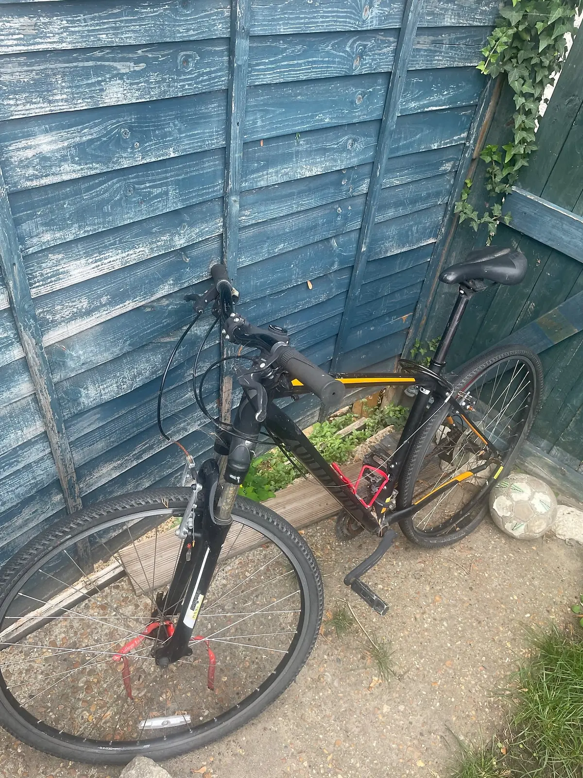 Black and yellow hybrid bike on stand, drive side, parked against weathered blue wooden fence in garden setting.
