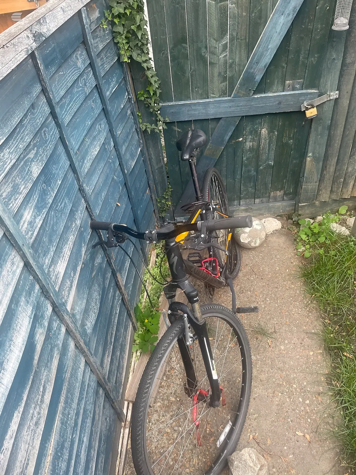 Black and yellow hybrid bike with red accents, parked against weathered blue-green wooden gates, non-drive side view, in a narrow alley.