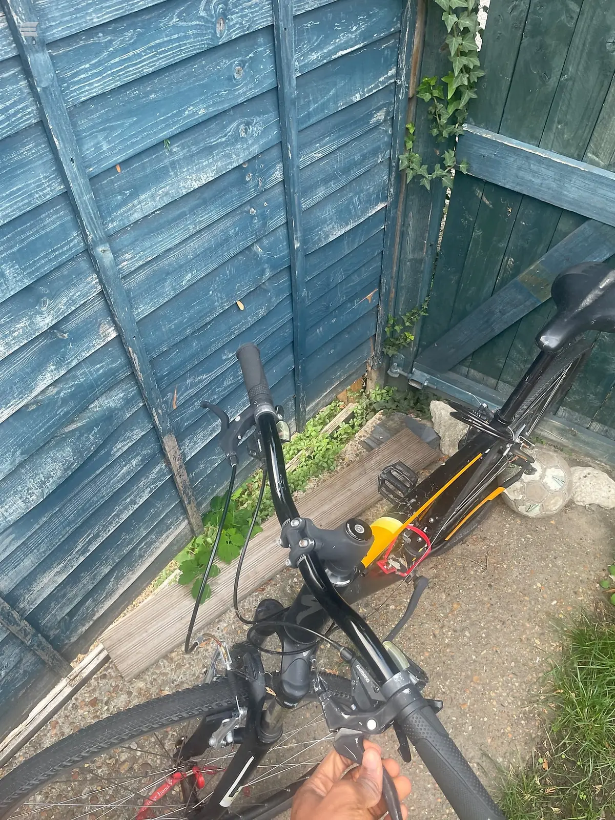 Black and yellow folding bike, three-quarter view from above, parked against weathered blue wooden sheds in a garden setting.