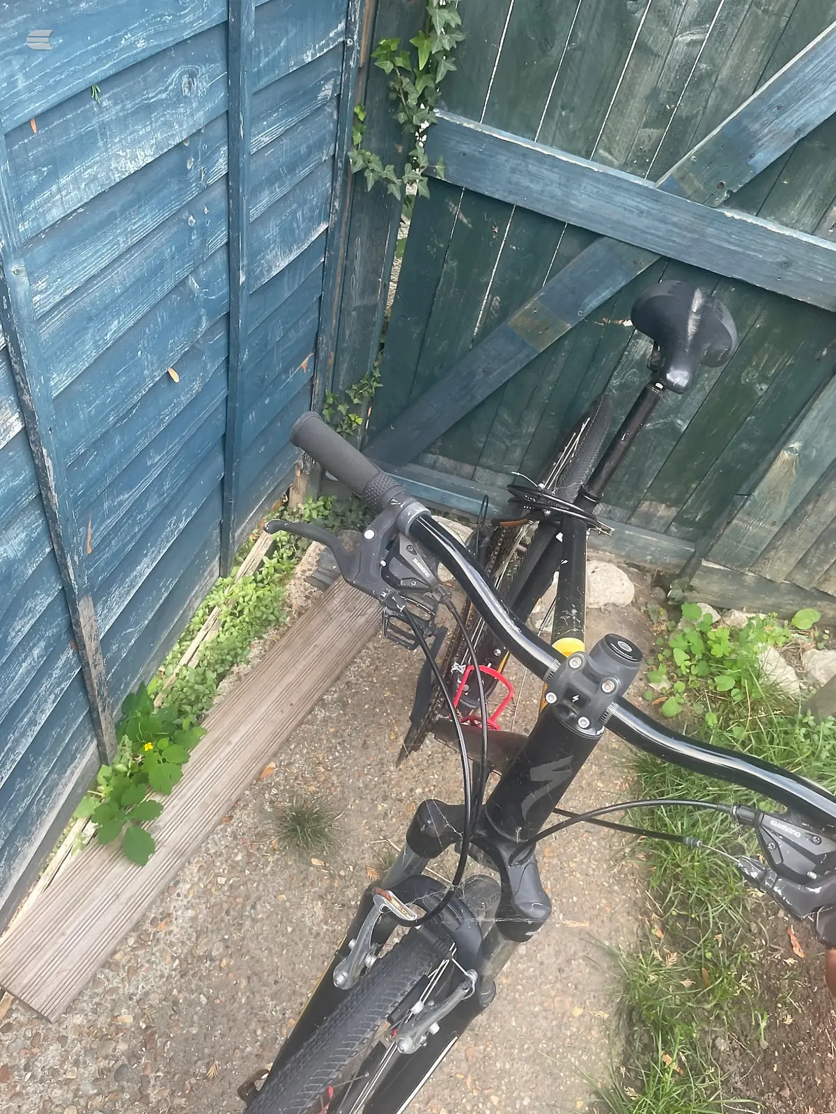 Black hybrid bike with drop bars, parked in a narrow alley between blue wooden sheds, non-drive side view, handlebars and both wheels visible.