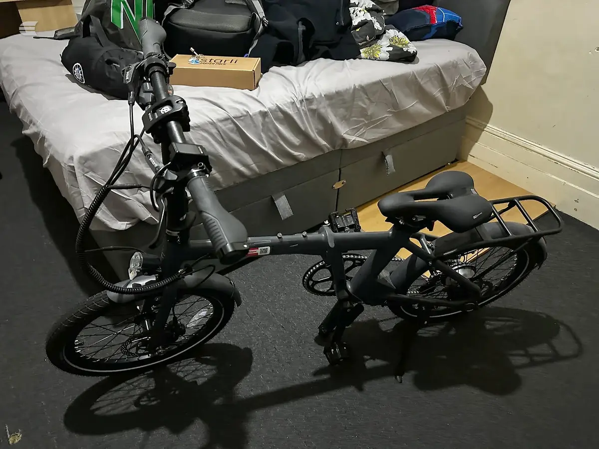 Black electric bike on dark floor indoors, non-drive side, with bed and storage visible behind.