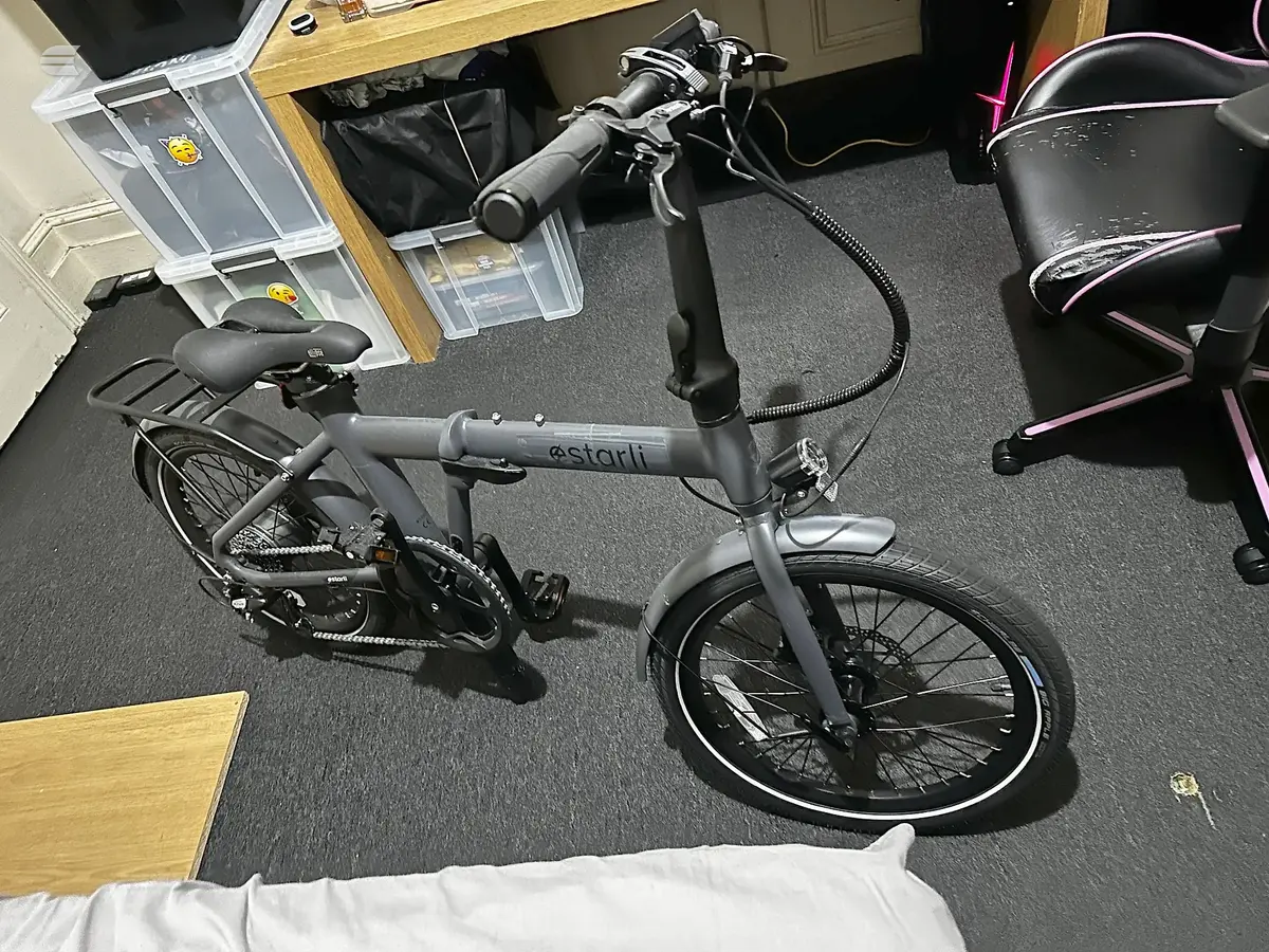 Grey Starli electric folding bike on dark carpet, three-quarter view from drive side, in an indoor office or workspace setting.