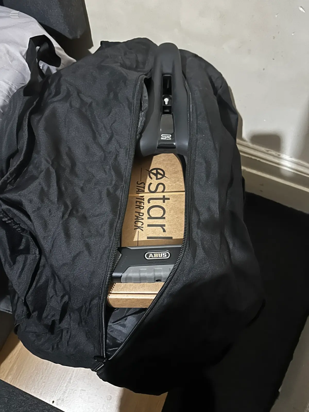 Abus Stelkoi smart lock with wooden packaging tag attached to black backpack, indoors against white wall.