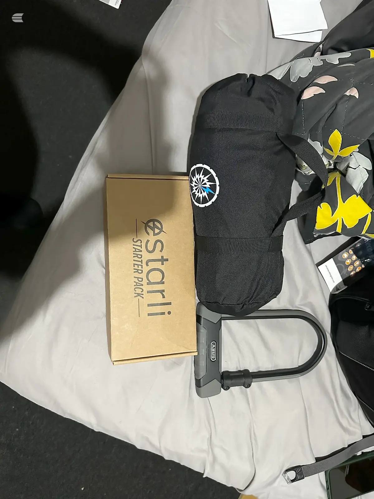 Estathé Starter Pack box with black insulated bottle bag featuring compass logo, U-lock, and floral-patterned black fabric item on white bedding.