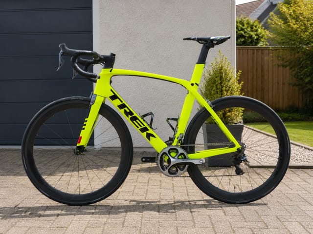 Neon yellow Trek road bike on a driveway, drive side view, against a modern house with dark garage door and wooden fence.
